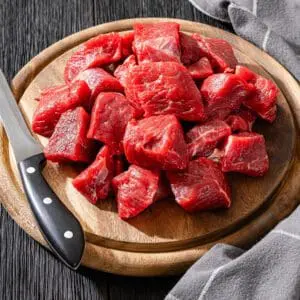 Raw beef cubes on wooden cutting board.